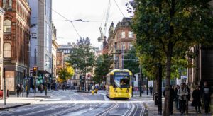 A photo of a Metrolink tram, part of the Manchester Bee Network, driving through Manchester city centre. 